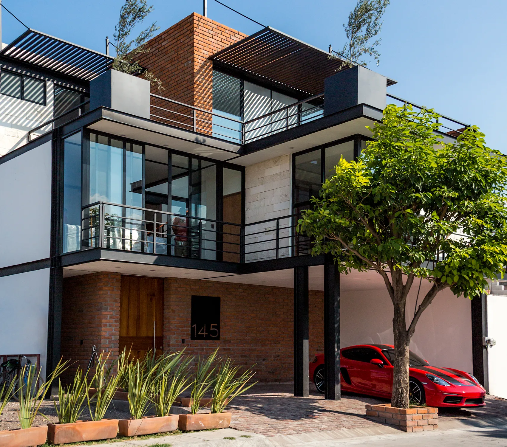 Fachada residencial estilo industrial moderno: estructura de acero negro (vigas IPR) enmarcando volúmenes de ladrillo rojo aparente y grandes ventanales de cristal en esquina.